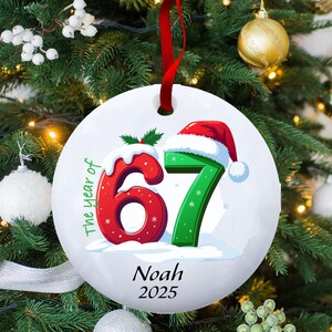 May include: White ceramic Christmas ornament with a red ribbon. The ornament features the numbers "67" in red and green, with a Santa hat on the "7". Text includes "The year of", "Noah", and "2025".