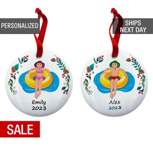 May include: Two white, round Christmas ornaments with red ribbon hangers. Each ornament features a cartoon person in a yellow inner tube in blue water, surrounded by a floral design. One says "Emily 2023", the other says "Alex 2023".