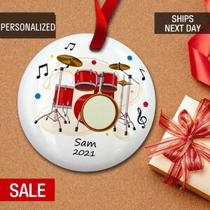 May include: A white ceramic Christmas ornament with a red drum set, musical notes, and the text "Sam 2021".