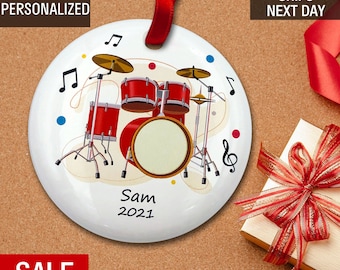 May include: A white ceramic Christmas ornament with a red drum set, musical notes, and the text "Sam 2021".