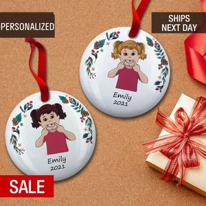 May include: Two white round ceramic ornaments with a red ribbon. Each ornament features a cartoon girl with brown hair and a red shirt, holding her fingers to her mouth. The ornaments have a green wreath with red berries and pine cones. The text "Emily 2021" is printed on each ornament.