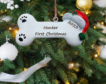May include: White bone-shaped Christmas ornament with paw prints and the text "Hunter First Christmas 2023" on a green Christmas tree.