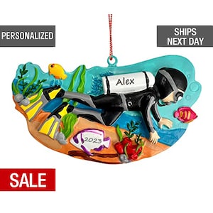 May include: A personalized Christmas ornament featuring a scuba diver with an oxygen tank, flippers, and a mask. The ornament is painted in shades of blue, green, and orange, with a white fish and coral reef details. The diver's tank has the name "Alex" written on it. The ornament also includes the year "2023" written on a purple fish.