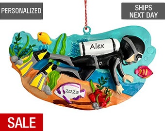May include: A personalized Christmas ornament featuring a scuba diver with an oxygen tank, flippers, and a mask. The ornament is painted in shades of blue, green, and orange, with a white fish and coral reef details. The diver's tank has the name "Alex" written on it. The ornament also includes the year "2023" written on a purple fish.
