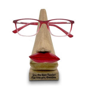 May include: Wooden eyeglasses holder shaped like a face with red lips and a nose. The holder has a message engraved on the base that reads "You Are Spec-Tacular! Eye love you, Grandma".