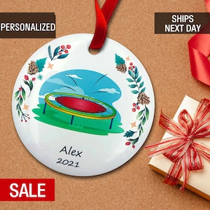 May include: White ceramic Christmas ornament with a colorful illustration of a trampoline and a green, red, and yellow border. The ornament is decorated with holly, pine cones, and stars. The text "Alex 2021" is written on the ornament.