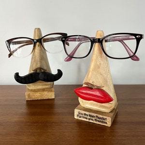 May include: Two wooden eyeglass holders shaped like faces. The holder on the left has a black mustache and the holder on the right has red lips and the text "You are Spec-tacular! Eye love you, Grandma".