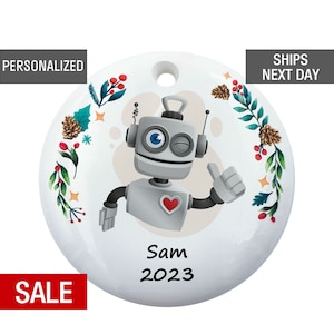 May include: White round Christmas ornament with a cartoon robot wearing a red heart. The robot is winking and giving a thumbs up. The ornament is decorated with green leaves, red berries, and pine cones. The text "Sam 2023" is printed on the ornament.