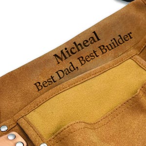 May include: Close-up of a brown leather tool belt with the engraved text "Micheal Best Dad, Best Builder". The tool belt has a light brown interior and metal rivets.
