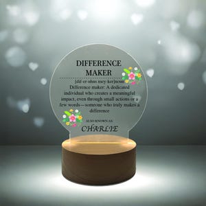 May include: A clear acrylic plaque with a wooden base and a light inside. The plaque has a floral design and the text "DIFFERENCE MAKER [dif-er-uhns mey-ker]noun Difference maker: A dedicated individual who creates a meaningful impact, even through small actions or a few words-someone who truly makes a difference ALSO KNOWN AS: CHARLIE"