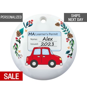 May include: White round ornament with a red car and a blue and white learner's permit with the text "MA Learner's Permit", "Name: Alex", and "Issued: 2023". The ornament is decorated with green leaves, red berries, and gold stars.