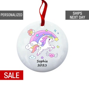 May include: White ceramic ornament with a rainbow, a unicorn, clouds, stars, and a heart. The ornament is personalized with the name "Sophia" and the year "2023".