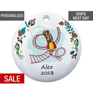 May include: White round Christmas ornament with a colorful illustration of a roller coaster with people riding it. The ornament has a green and red floral border and the text "Alex 2023" in black.