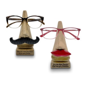 May include: Two wooden eyeglass holders shaped like faces. The holder on the left has a mustache and the text "They are here Grandpal Love you, Emma". The holder on the right has red lips and the text "You Are Spee-Tacular! Eye love you, Grandma".