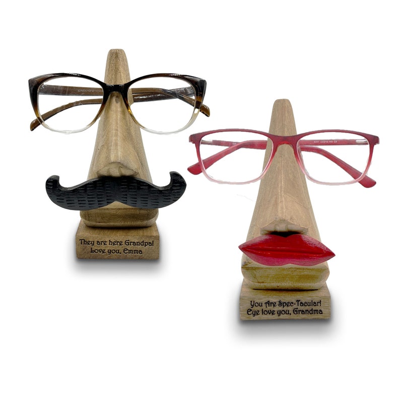 May include: Two wooden eyeglass holders shaped like faces. The holder on the left has a mustache and the text "They are here Grandpal Love you, Emma". The holder on the right has red lips and the text "You Are Spee-Tacular! Eye love you, Grandma".