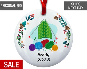 May include: A white round ceramic ornament with a green knitted sweater, colorful yarn balls, and knitting needles. The ornament has a blue background and a floral wreath around the edge. The text "Emily 2023" is printed on the ornament.