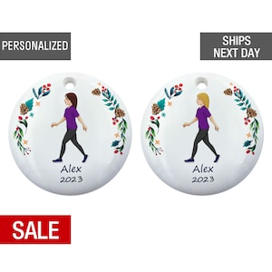 May include: A white round Christmas ornament with a green, red, and gold floral wreath around the edge. A cartoon illustration of a person walking is in the center of the ornament. The text "Alex 2023" is below the person.