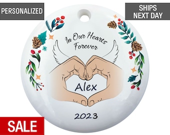 May include: White round ceramic ornament with a hand-drawn design of two hands forming a heart shape with the name "Alex" inside. The ornament has a green and red floral border with the text "In Our Hearts Forever" and the year "2023".