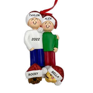 May include: Personalized Christmas ornament featuring a family of four with two dogs. The ornament is white with red and green accents. The family members are wearing Santa hats with their names on them: Taylor, Alex, Rocky, and Bella. The year 2022 is written on the ornament.