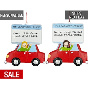 May include: Red car ornament with a learner's permit sign on top. The sign reads "NC LEARNER'S PERMIT" with the name "Sofia Green" and the date "09.09.2022". The second ornament is a red car with a learner's permit sign that reads "NY LEARNER'S PERMIT" with the name "Nicky Parsons" and the date "04/16/2022".