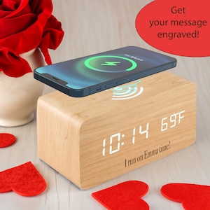 May include: A wooden alarm clock with a digital display showing the time 10:14 and the temperature 69°F. The clock has a built-in wireless charging pad with a smartphone on top. The clock has a personalized message engraved on the front: "I run on Emma time!"
