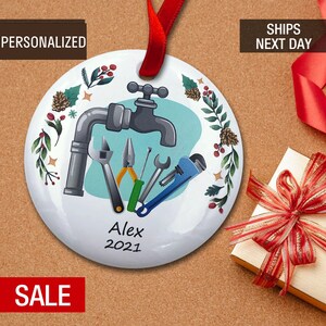 May include: A white round Christmas ornament with a blue background featuring a cartoon illustration of a faucet and various plumbing tools. The ornament is decorated with green leaves, red berries, and pine cones. The text "Alex 2021" is printed on the ornament.