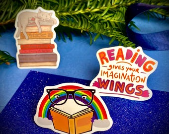 Op de afbeelding: Drie kleurrijke stickers met een lees-thema. Eén sticker toont een kat die slaapt op een stapel boeken. Een andere sticker toont een boek met een regenboog en een bril. De derde sticker zegt "Lezen geeft je verbeelding vleugels".