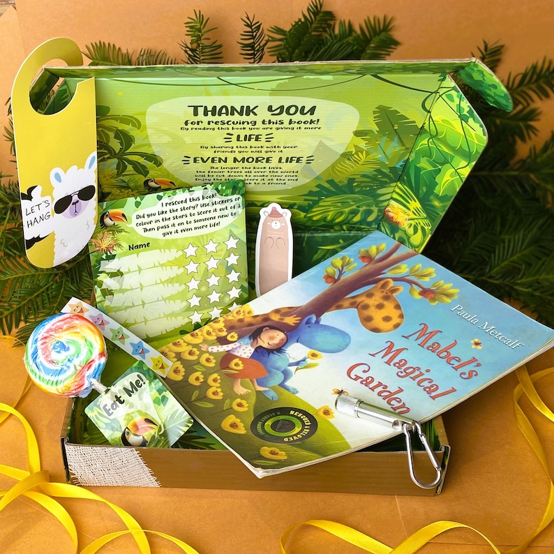May include: A green and brown cardboard box with a book titled "Mabel's Magical Garden" by Paula Metcalf. The box also contains a lollipop, a sticker sheet, a bookmark, and a small keychain. The box has a thank you message for rescuing the book and a message about earning more life.