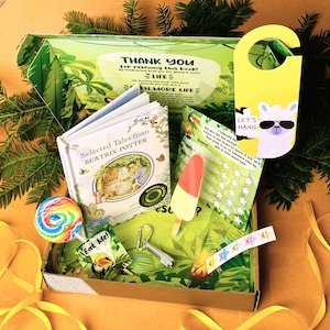 May include: A brown cardboard box with a green and white jungle-themed design. The box is open and contains a variety of items, including a book of Beatrix Potter stories, a lollipop, a flashlight, a popsicle, a llama door hanger, and a sticker sheet. The box is surrounded by yellow ribbon.