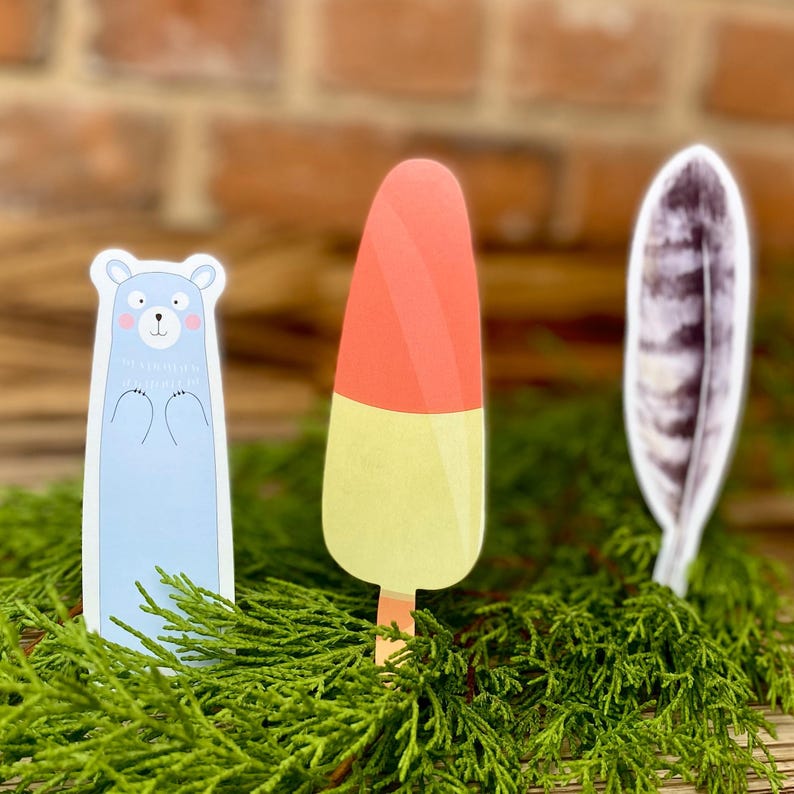 May include: Three paper cutouts of a bear, an ice cream cone, and a feather. The bear is light blue with a pink nose and is standing upright. The ice cream cone is red and yellow and is standing upright. The feather is gray and white and is lying on its side. All three cutouts are on a bed of green foliage.
