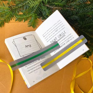 May include: A close-up of a book with a grey and green bookmark. The bookmark has the text "Tight Circle" and "Information Leaks" printed on it. The book has the text "Amy Knows!" printed on it.