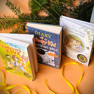 May include: Three paperback books on a brown surface with yellow ribbon. The books are "Diary of a Wimpy Kid", "Five Go Off in a Caravan", and "Selected Tales from Beatrix Potter".