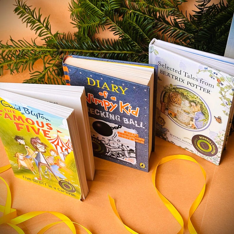 May include: Three paperback books on a brown surface with yellow ribbon. The books are "Diary of a Wimpy Kid", "Five Go Off in a Caravan", and "Selected Tales from Beatrix Potter".