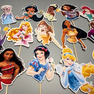 May include: A set of 12 princess themed cake toppers. The toppers feature various Disney princesses, including Snow White, Cinderella, Ariel, Belle, and Mulan. Each topper is printed on cardstock and has a wooden stick attached to the back.