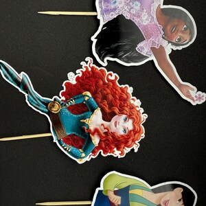 May include: Three princess cake toppers featuring Merida with red hair, Mulan with black hair, and Tiana with brown hair. Each topper is printed on cardstock and attached to a wooden stick.