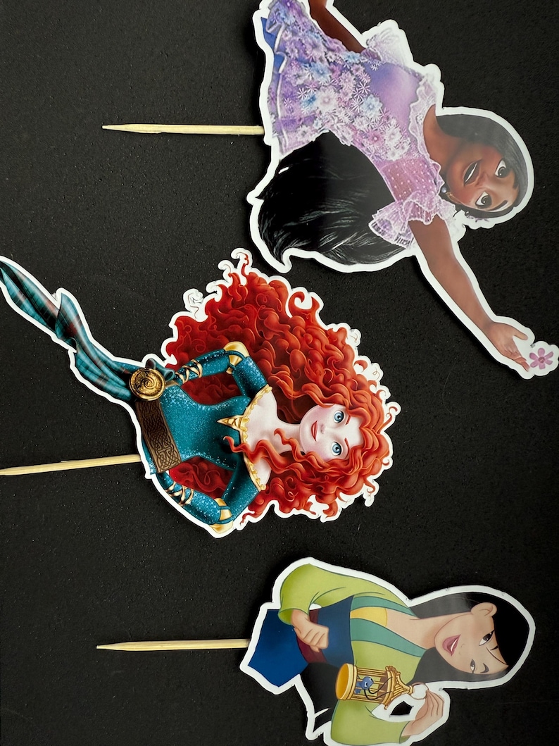 May include: Three princess cake toppers featuring Merida with red hair, Mulan with black hair, and Tiana with brown hair. Each topper is printed on cardstock and attached to a wooden stick.