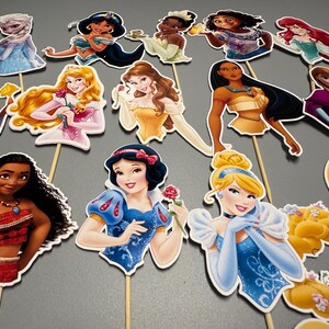 May include: A set of 14 princess character cake toppers. The toppers are printed on cardstock and feature various Disney princesses, including Snow White, Cinderella, Ariel, Belle, and Moana. Each topper has a stick attached to the back for easy insertion into a cake.