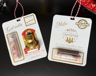 May include: Two white gift tags with gold lettering and red and white string. One tag says "Gabriella" and features a teddy bear and a roll of paper. The other says "Milo" and "Merry Christmas" with a roll of paper.