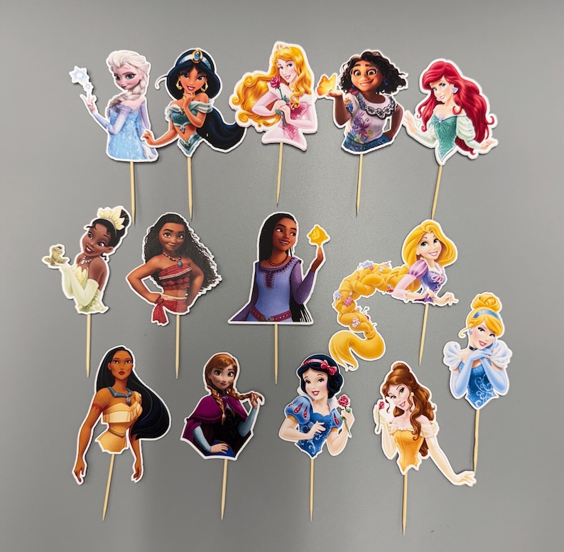 May include: Set of 12 princess themed cake toppers. Each topper features a different Disney princess character, including Ariel, Belle, Cinderella, Elsa, Jasmine, Mulan, Pocahontas, Rapunzel, Snow White, Tiana, and Moana. The toppers are printed on cardstock and attached to wooden skewers.