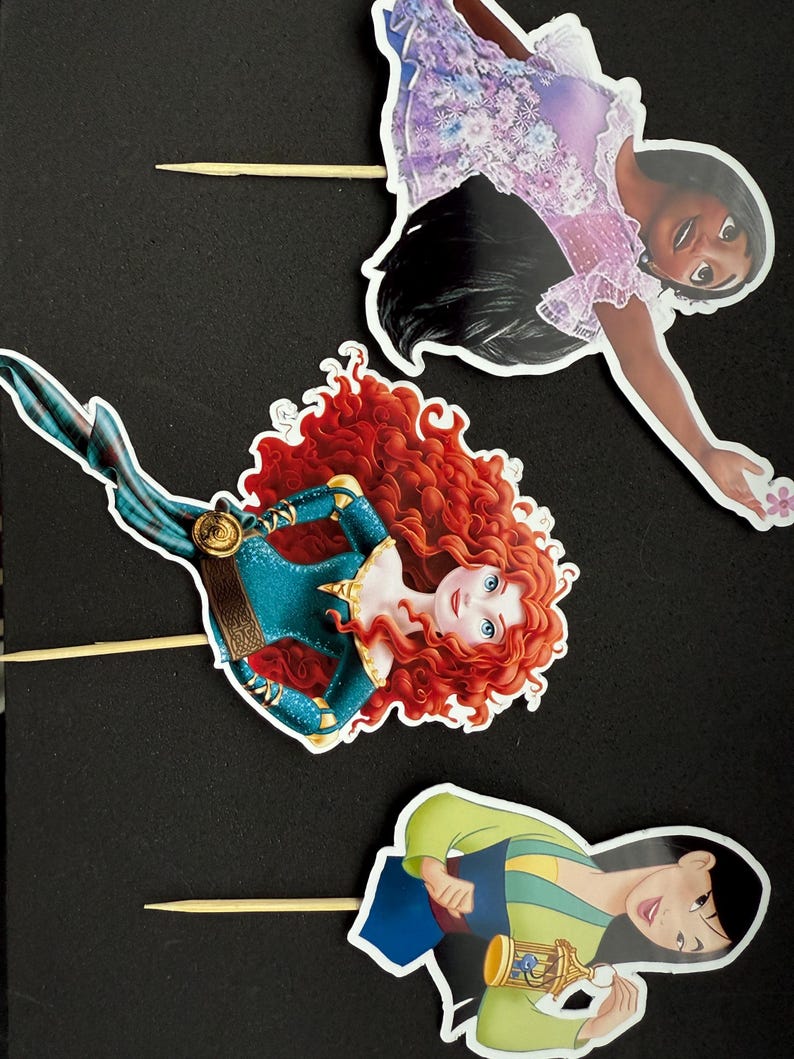 May include: Three princess cake toppers featuring Mulan, Merida, and Tiana. The toppers are printed on cardstock and have a stick attached to the back.