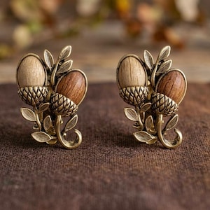 May include: A pair of acorn-shaped earrings. Each earring features two acorns, one light-colored and one dark-colored, set in a gold-toned metal with leaf accents. The earrings are displayed on a brown fabric surface.