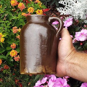May include: A dark brown ceramic jug with a smooth, glossy finish and a single side handle. The vessel features a cylindrical body with a slightly tapered neck and a wide opening. The base shows a lighter tan color where the glaze is thinner.