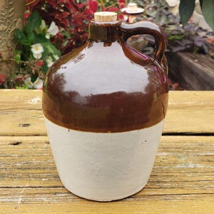 May include: A ceramic jug featuring a two-tone design with a glossy dark brown glazed upper section and a matte off-white lower section. The jug has a single side handle, a narrow neck, and a small cork stopper inserted into the opening.