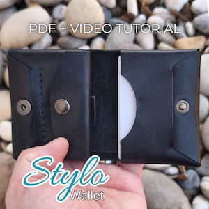 May include: Black leather wallet with a snap closure, open to reveal a white card. The text "PDF + VIDEO TUTORIAL" is at the top, and "Stylo Wallet" is at the bottom. The wallet is on a background of small, smooth stones.