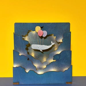 Könnte beinhalten: Ein Holzwal mit drei Ballons fliegt durch einen blauen Himmel mit weißen Wolken. Der Wal und die Ballons sind aus dem Holz ausgeschnitten und der Himmel ist blau bemalt. Das Stück wird von hinten mit weißen Lichtern beleuchtet.