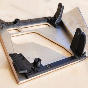 Puede incluir: Un componente negro impreso en 3D con una base rectangular y soportes angulados, fijado a una superficie de madera clara. El componente tiene múltiples orificios para tornillos y un diseño recortado. La estructura es un soporte de montaje.