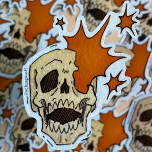 May include: A sticker featuring a skull with an orange burst design. The skull is beige with black eye sockets and a wide open mouth. The sticker has a white border and the text "@sebaspool5".