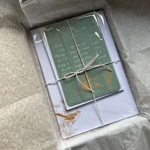 May include: A gift set in a white box, featuring a stack of green bookmarks tied with twine, and a white card with a bird illustration. The bookmarks have text and illustrations. The box is lined with white tissue paper.