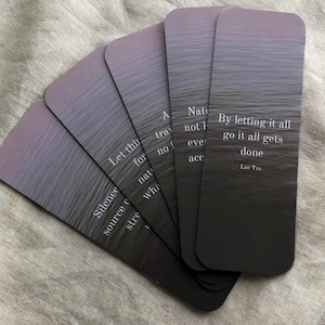May include: Five rectangular cards with a grey and purple gradient design are fanned out. Each card features a quote in white text. The quotes include: "By letting it all go it all gets done" and "Silence source of strength".