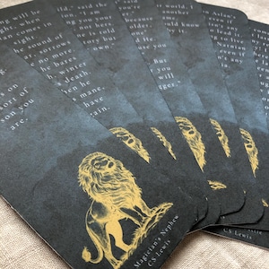 May include: A collection of dark grey bookmarks with gold text and illustrations. Each bookmark features a quote from "The Magician's Nephew" by C.S. Lewis, with a gold lion illustration. The bookmarks are fanned out.